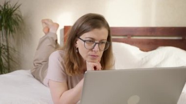 A young woman uses a laptop while lying on the bed in the room.