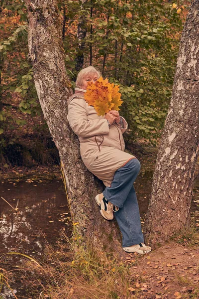 一位穿着浅色夹克的白发苍苍的老妇人 提着一束树叶在公园里散步 — 图库照片