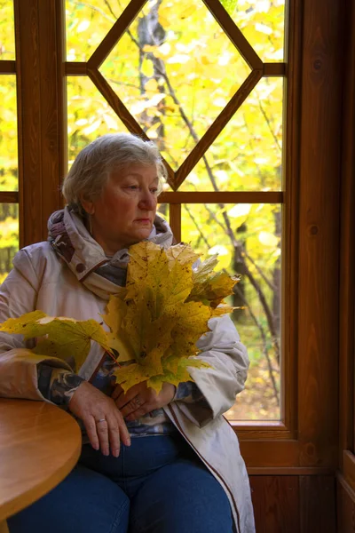 一个悲伤的 灰白头发的 65岁的女人坐在窗边阳台上的画像 在秋树的背景下 — 图库照片