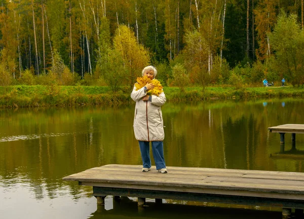 秋天公园的一座池塘上 站在一座木桥上的一位老妇人站着一束黄色枫叶 穿着一件浅薄的夹克 — 图库照片