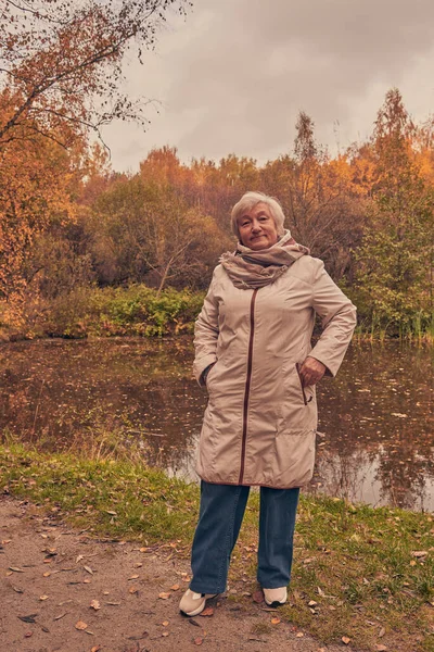 An elderly woman stands on the shore of a pond on a cloudy autumn day. — Stock Photo, Image