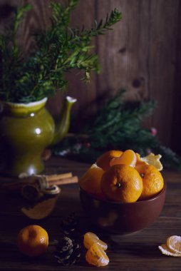 Kahverengi bir seramik kasede taze taze mandalinalar yatıyor. Arka planda dallar.