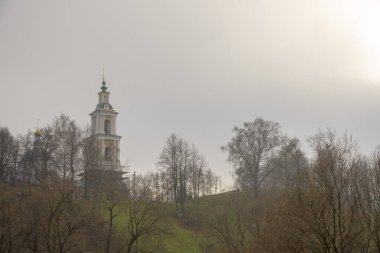 Tepede bulutlu bir gökyüzüne karşı uzun bir çan kulesi var. Güneş bulutların arasından bakıyor. Puslu kırsal sonbahar manzarası.