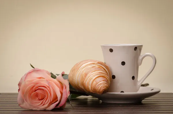 Croissant, šálek s podšálkem a růžové růže — Stock fotografie