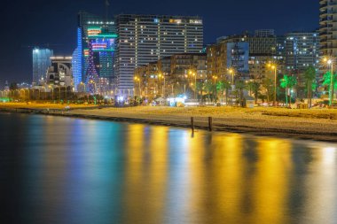 Deniz yüzeyine yansıyan modern mimari ışıklarla aydınlatılmış bir gece plajı, deniz manzarası.