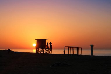 Altın günbatımı sahilinde bir cankurtaran kulesi silueti.