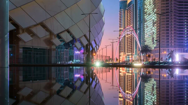 View of the colorful illuminated stadium and night city with modern architecture reflecting on the water surface