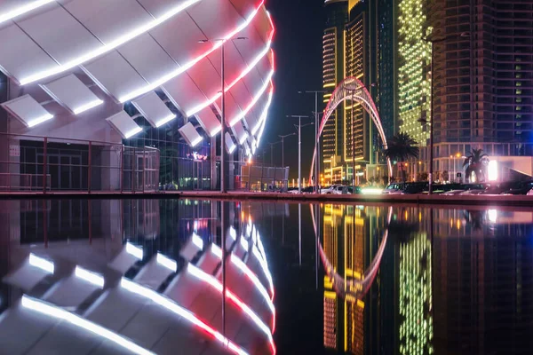 View of the colorful illuminated stadium and night city with modern architecture reflecting on the water surface