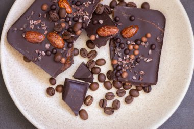 Chopped dark chocolate with almond and coffee beans. Broken chocolate on beige plate. Sweet food. Dark chocolate with nuts. Coffee product. 