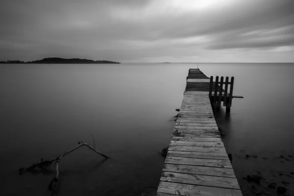 Long exposure view of a pier on a lake with perfectly still water beneath a dramatic, moody sky — Stock Photo, Image