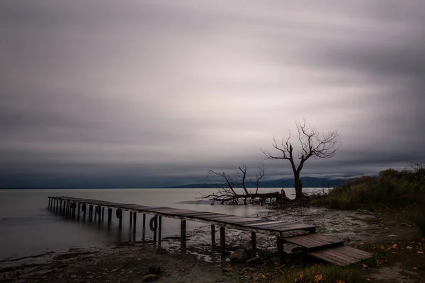 Veduta di un molo su un lago, sotto un cielo drammatico e lunatico con alberi scheletrici e spogli sullo sfondo — Foto Stock