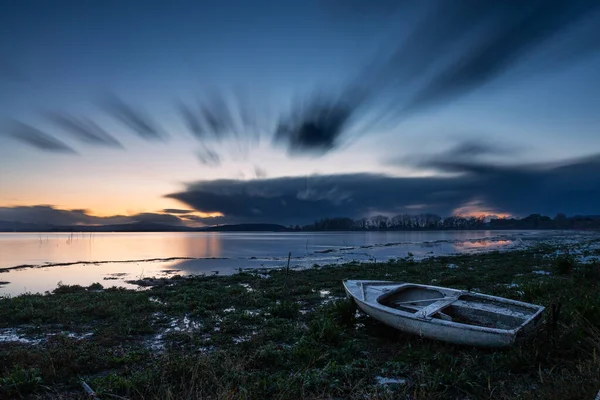 Long exposure view of shore of Trasimeno lake Umbria, Italy with a little boat at dusk, with perfectly still water and moving clouds.