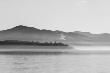 Alacakaranlıktaki Trasimeno Gölü 'nün huzurlu manzarası, sislerin arasından görünen ada ve tepeler..