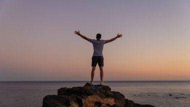 Arkaya bakan adam zafer pozu veriyordu. Esmer erkek el ele tutuşarak gökyüzüne yükseliyor. Tenerife Adası 'ndaki okyanusa bakıyor. İnsan ve doğa kavramı.