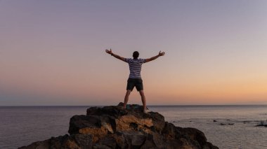 Arkaya bakan adam zafer pozu veriyordu. Esmer erkek el ele tutuşarak gökyüzüne yükseliyor. Tenerife Adası 'ndaki okyanusa bakıyor. İnsan ve doğa kavramı.