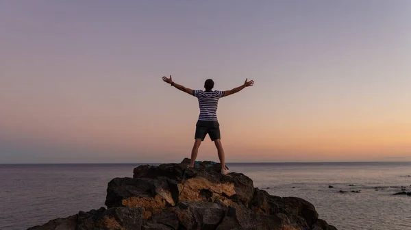 Arkaya bakan adam zafer pozu veriyordu. Esmer erkek el ele tutuşarak gökyüzüne yükseliyor. Tenerife Adası 'ndaki okyanusa bakıyor. İnsan ve doğa kavramı.