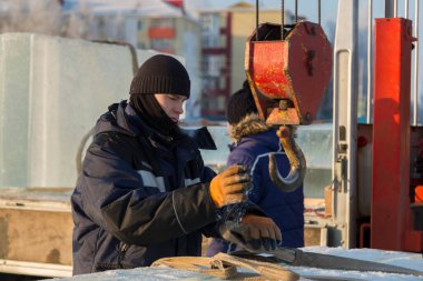 Mavi ceketli, kapüşonlu bir işçinin portresi. Buz kasabası inşaatında bir vinç kancasına takılmış.