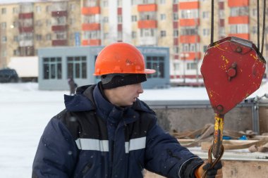 Mavi ceketli, kapüşonlu bir işçinin portresi. Buz kasabası inşaatında bir vinç kancasına takılmış.