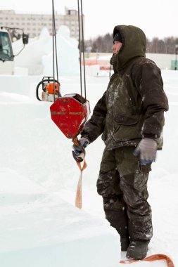 Kapüşonlu yeşil ceketli bir işçinin portresi. Buz kalıplarını yükleyen bir kamyonun kancasına takılmış.