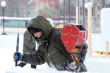 Kapüşonlu yeşil ceketli bir işçinin portresi. Buz kalıplarını yükleyen bir kamyonun kancasına takılmış.