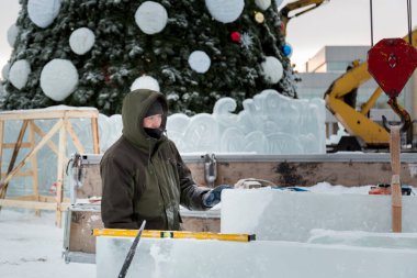 Kapüşonlu yeşil ceketli bir işçinin portresi. Buz kalıplarını yükleyen bir kamyonun kancasına takılmış.