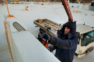 Kafasında kapüşonlu mavi ceketi olan işçi bir tesisatçı elektrikli testere ile bir buz plakası kullanıyor.