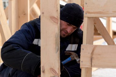 Kulaklıklı bir şapka takan biri ahşap kirişlerden yapılmış bir kaydırağın çerçevesini monte eder.