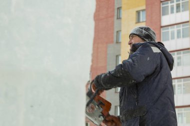 Mavi ceketli bir montaj işçisi ve siyah örülmüş bir şapka bir buz tabakasını testere ile düzeltiyor.