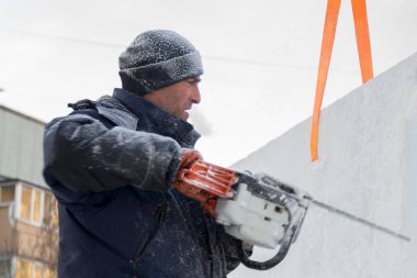 Mavi ceketli bir montaj işçisi ve siyah örülmüş bir şapka bir buz tabakasını testere ile düzeltiyor.