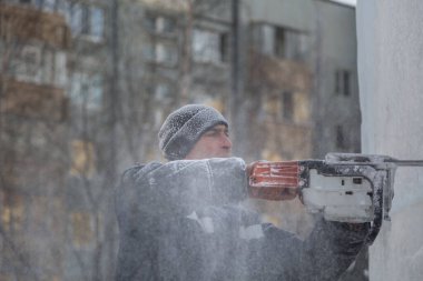 Mavi ceketli bir montaj işçisi ve siyah örülmüş bir şapka bir buz tabakasını testere ile düzeltiyor.