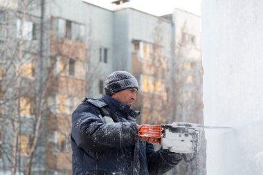 Mavi ceketli bir montaj işçisi ve siyah örülmüş bir şapka bir buz tabakasını testere ile düzeltiyor.