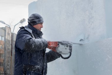 Mavi ceketli bir montaj işçisi ve siyah örülmüş bir şapka bir buz tabakasını testere ile düzeltiyor.
