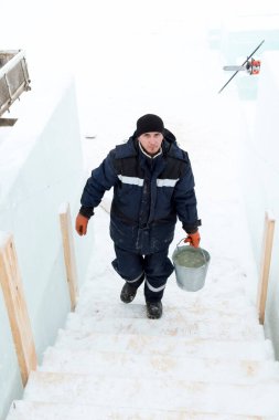Mavi ceketli ve siyah şapkalı bir işçi elinde bir kova karla merdivenlerden yukarı çıkıyor.