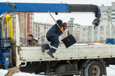 Kapüşonlu ceketli ve kafasında kırmızı örgü şapkalı bir işçi elinde kürekle arabanın arkasından atlıyor.