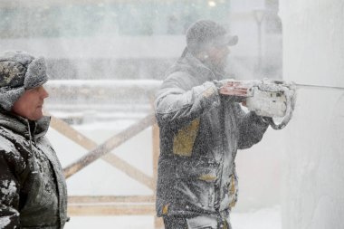 İnşaat alanında çalışan iki işçi, bir benzin testeresiyle buz paneline zarif bir Noel kompozisyonu oyuyorlar..