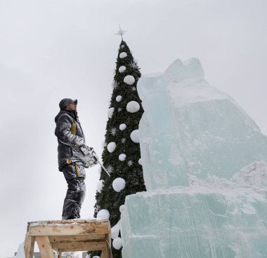 Heykeltıraş, buz kalıbından bir buz kalıbını bir yılbaşı ağacının arka planına elektrikli testere ile keser..