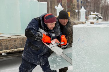 Mavi ceketli işçi derleyicisi ve örgülü bir şapka bir buz tabakasını testereyle kesiyor.