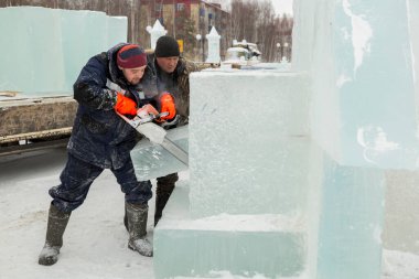 Mavi ceketli işçi derleyicisi ve örgülü bir şapka bir buz tabakasını testereyle kesiyor.