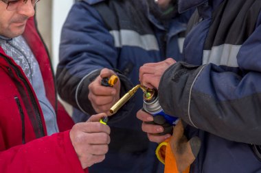 İnşaat alanında fermuarı açık kırmızı ceketli genç bir adam gaz ocağını yakıyor.