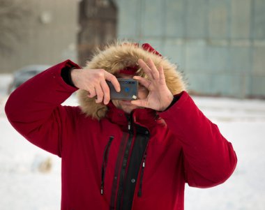 Kışın kırmızı ceketli, gözlüklü genç bir adam cep telefonuyla fotoğraf çekiyor.