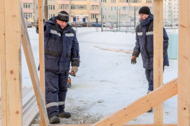Elinde çekiç, elinde mavi bir ceketle ahşap bir binanın kaputu olan bir inşaatçı.