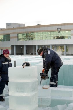 Mavi ceketli bir montaj işçisi ve siyah örülmüş bir şapka bir buz tabakasını testere ile düzeltiyor.