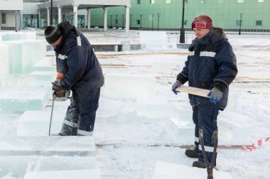 Mavi ceketli bir montaj işçisi ve siyah örülmüş bir şapka bir buz tabakasını testere ile düzeltiyor.