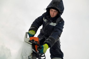 Kafasında kapüşonlu mavi ceketi olan işçi bir tesisatçı elektrikli testere ile bir buz plakası kullanıyor.