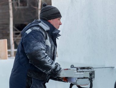 Mavi ceketli ve örgü şapkalı çalışan bir derleyici elektrikli testere ile buz kalıbı sürüyor.