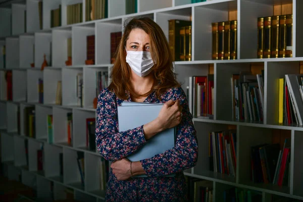 Young woman with mask for corona virus holding big books in library. Business girl at the office