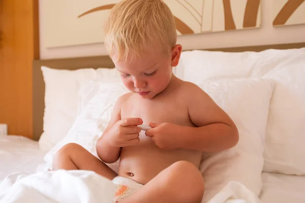 A little boy sits in bed and holds a thermometer in his hands and looks at his temperature