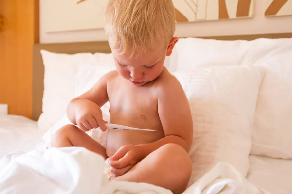 A little boy sits in bed and holds a thermometer in his hands and looks at his temperature
