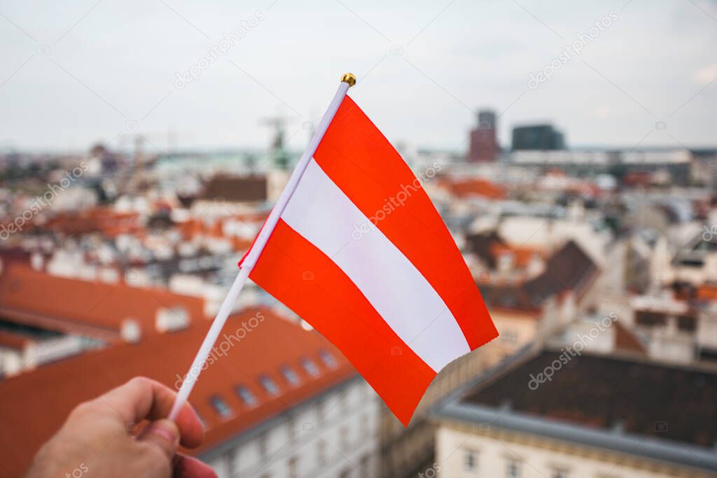 A man holds in his hand the flag of Austria on a viewing platform of St. Stephens Cathedral in Vienna against the backdrop of a beautiful sunset sky and city panorama.