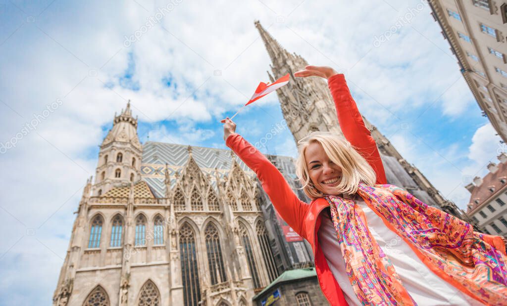 Young beautiful smiling woman in a red jacket with the flag of Austria in hand stands on the background of St. Stephens Cathedral in Vienna , Austria